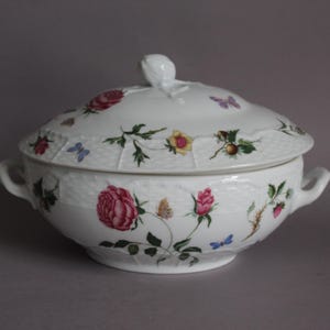 Könnte beinhalten: Weißes Porzellan-Serviergeschirr mit Deckel, verziert mit rosa Rosen, gelben Blumen, grünen Blättern und blauen Schmetterlingen. Das Geschirr hat zwei Griffe und einen dekorativen Deckel mit floralem Knauf. Ein Klassiker.