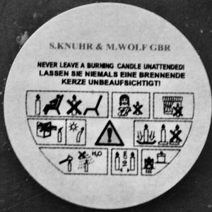 May include: Black and white warning label with the text "S.KNUHR & M.WOLF GBR" and "NEVER LEAVE A BURNING CANDLE UNATTENDED! LASSEN SIE NIEMALS EINE BRENNENDE KERZE UNBEAUFSICHTIGTI" along with several safety pictograms.
