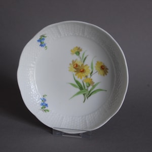 Könnte beinhalten: Weißer Keramikteller mit strukturiertem Rand und floralem Design. Der Teller zeigt gelbe Blumen mit grünen Blättern in der Mitte und kleine blaue Blumen an der Seite. Der Teller steht auf einem transparenten Ständer.