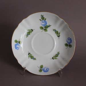 May include: White saucer with a scalloped edge and gold trim. The saucer is decorated with blue rose and green leaf designs. The saucer is displayed on a clear stand.