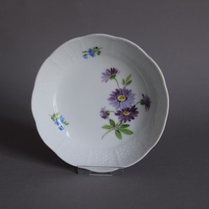 May include: White ceramic plate with a scalloped edge and a floral design. The plate features a cluster of purple and yellow flowers with green leaves, and small blue flowers. The plate has a textured surface.