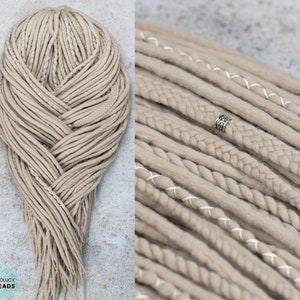 Pu&ograve; includere: Un set di dreadlock sintetici color beige con un mix di ciocche lisce, intrecciate e cucite con filo bianco. La collezione include una perlina in metallo argentato con incisioni complesse. Il testo BOHOLUCY DREADS &egrave; visibile in basso.