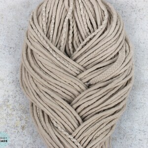 Pu&ograve; includere: Un set di dreadlocks sintetici color beige disposti in un fascio intrecciato spesso. Le ciocche presentano texture varie, tra cui tubi lisci, sezioni intrecciate e cuciture decorative bianche, con un piccolo dettaglio a perla in metallo argentato.