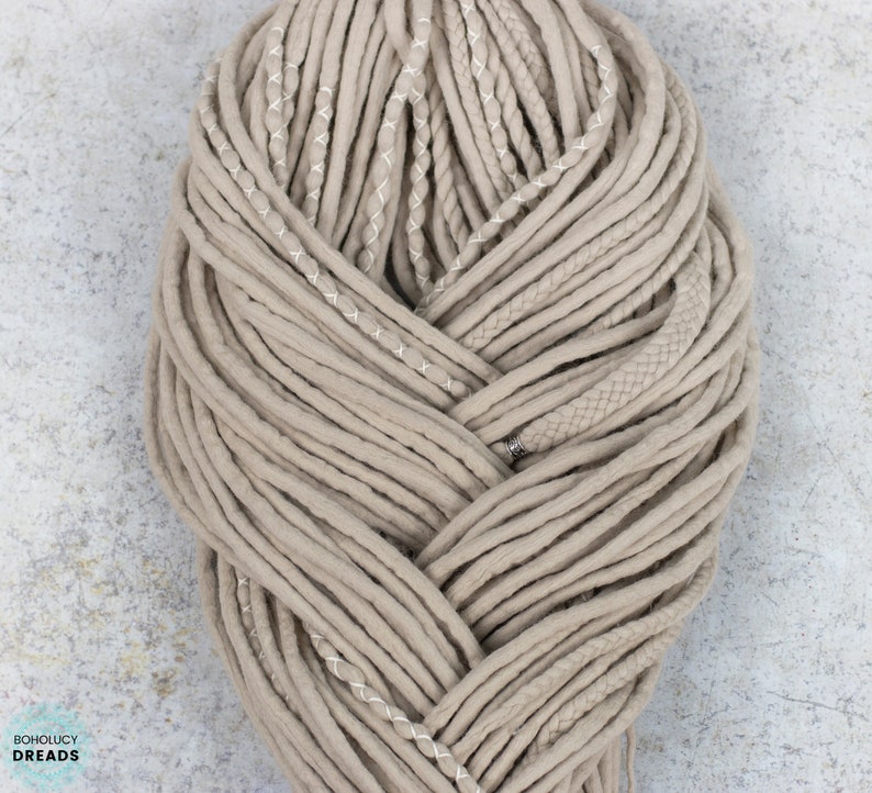 Pu&ograve; includere: Un set di dreadlocks sintetici color beige disposti in un fascio intrecciato spesso. Le ciocche presentano texture varie, tra cui tubi lisci, sezioni intrecciate e cuciture decorative bianche, con un piccolo dettaglio a perla in metallo argentato.