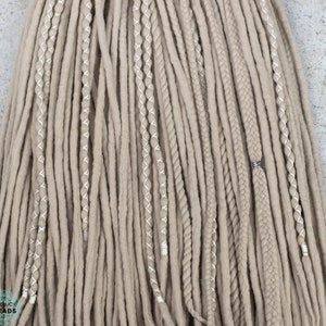 Pu&ograve; includere: Un set di dreadlock sintetici color beige caratterizzato da un mix di ciocche lisce, sezioni ritorte avvolte con filo bianco e trecce spesse. Una treccia &egrave; decorata con una piccola perlina in metallo color argento. Il testo BOHOLUCY DREADS &egrave; visibile.