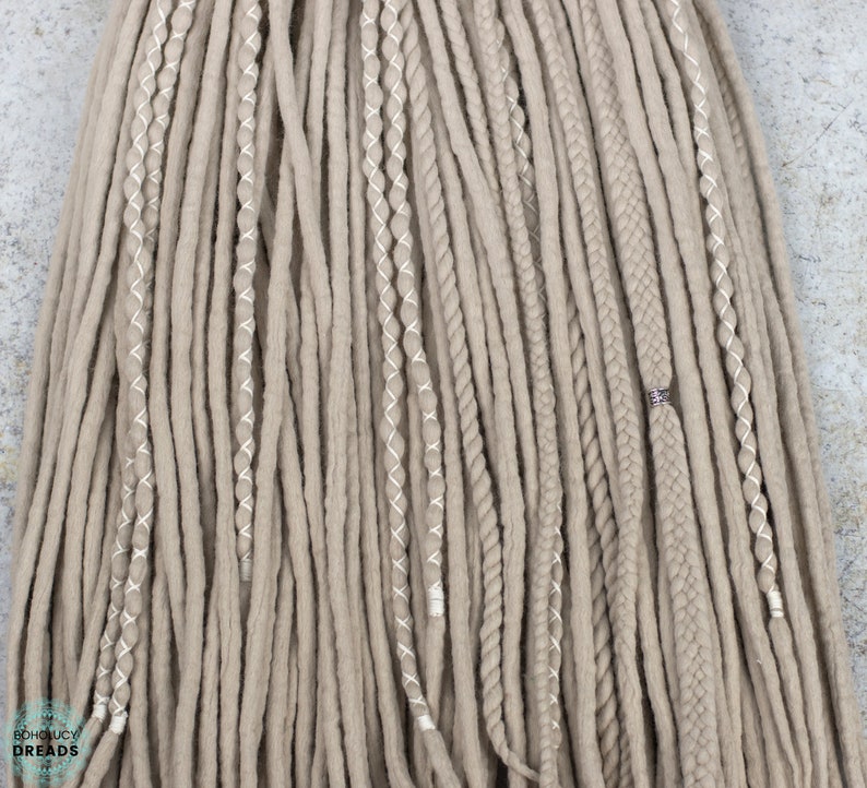 Pu&ograve; includere: Un set di dreadlock sintetici color beige caratterizzato da un mix di ciocche lisce, sezioni ritorte avvolte con filo bianco e trecce spesse. Una treccia &egrave; decorata con una piccola perlina in metallo color argento. Il testo BOHOLUCY DREADS &egrave; visibile.
