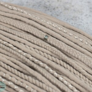 Pu&ograve; includere: Una collezione di dreadlocks sintetici color beige con varie texture, tra cui ciocche lisce, cordoncini intrecciati e ciocche avvolte con filo bianco. Una singola perlina in metallo color argento &egrave; fissata a uno dei dreadlocks centrali. Il testo BOHOLUCY DREADS &egrave; visibile nell'angolo.