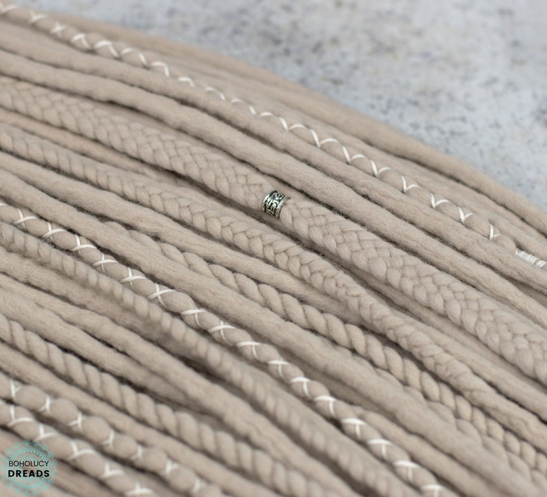 Pu&ograve; includere: Una collezione di dreadlocks sintetici color beige con varie texture, tra cui ciocche lisce, cordoncini intrecciati e ciocche avvolte con filo bianco. Una singola perlina in metallo color argento &egrave; fissata a uno dei dreadlocks centrali. Il testo BOHOLUCY DREADS &egrave; visibile nell'angolo.
