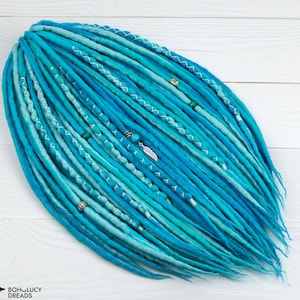 May include: A set of blue and turquoise synthetic dreadlocks with white accents and silver beads. The dreadlocks are arranged in a fan shape and are ready to be worn.