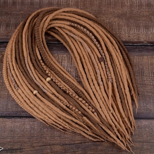 May include: A collection of handmade dreadlocks in shades of brown and tan. The dreads are arranged in a pile, displaying their texture and colour variations. Some dreads have decorative elements such as braids and wooden beads. The background is a dark wood surface.