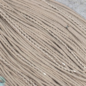 Pu&ograve; includere: Una collezione di dreadlocks sintetici color beige caratterizzata da un mix di ciocche lisce, texture intrecciate e ciocche avvolte con filo bianco. Una piccola perlina in metallo color argento &egrave; fissata su una treccia. Il testo Bohology Dreads &egrave; visibile.