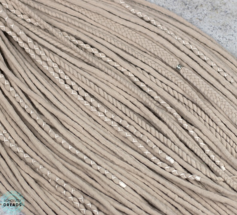 Pu&ograve; includere: Una collezione di dreadlocks sintetici color beige caratterizzata da un mix di ciocche lisce, texture intrecciate e ciocche avvolte con filo bianco. Una piccola perlina in metallo color argento &egrave; fissata su una treccia. Il testo Bohology Dreads &egrave; visibile.