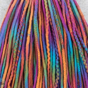 May include: Close-up of colorful dreadlocks in shades of purple, orange, blue, and green. Some dreads are accented with orange stitching or silver beads. The dreadlocks are arranged closely together, creating a vibrant and textured appearance.