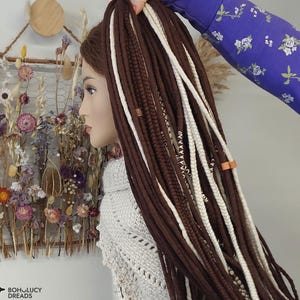 Puede incluir: Una cabeza de maniquí con largas rastas marrones oscuras y blancas. Las rastas están peinadas con una mezcla de mechones marrones y blancos, y están adornadas con pequeñas cuentas de madera. El maniquí lleva un suéter gris claro.