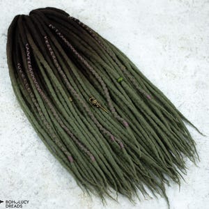 May include: Olive green and brown dreadlocks with decorative accents. The dreads are arranged in a layered pattern, with a mix of solid and textured sections. A small, ornate charm is attached to the dreads.