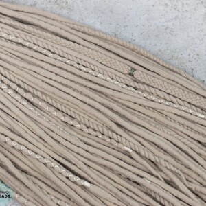 Pu&ograve; includere: Una collezione di dreadlocks sintetici color beige con varie texture, tra cui ciocche lisce, intrecciate e attorcigliate. Alcune ciocche sono avvolte da filo bianco o decorate con una piccola perlina in metallo argentato. Il testo BOHOLUCY DREADS &egrave; visibile.