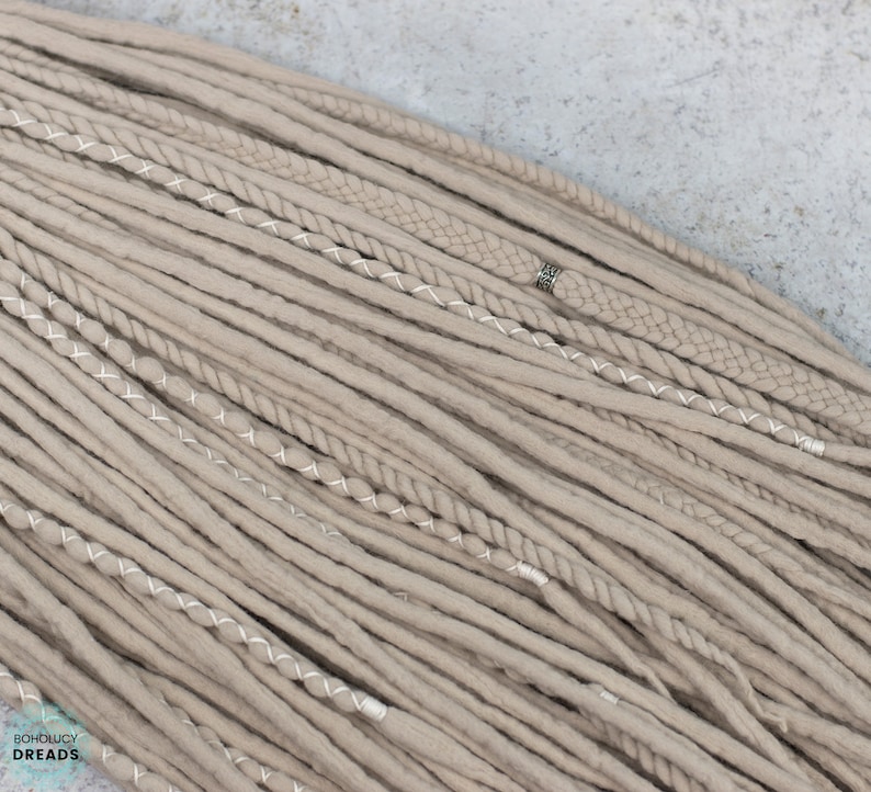 Pu&ograve; includere: Una collezione di dreadlocks sintetici color beige con varie texture, tra cui ciocche lisce, intrecciate e attorcigliate. Alcune ciocche sono avvolte da filo bianco o decorate con una piccola perlina in metallo argentato. Il testo BOHOLUCY DREADS &egrave; visibile.