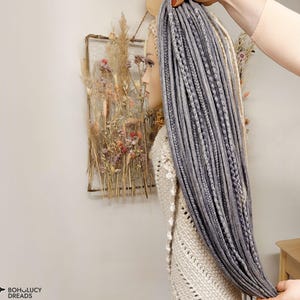 May include: A set of long synthetic dreadlocks in shades of grey and light blonde, featuring a mix of smooth and braided textures. The hair extensions are displayed on a mannequin head, showcasing a layered, multi-tonal appearance with various thicknesses.