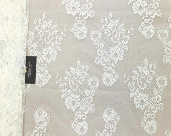May include: White lace fabric with a floral pattern. The lace features a delicate design of clustered flowers and leaves, with a scalloped edge. The fabric is semi-transparent, revealing a light-colored background. A black tag with white text is visible.