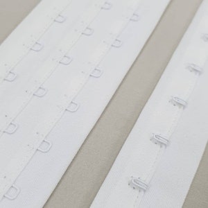 Può includere: Primo piano di chiusure per reggiseno bianche con ganci e occhielli. Le chiusure sono realizzate in tessuto bianco con più file di chiusure con ganci e occhielli. Lo sfondo è di colore beige neutro.
