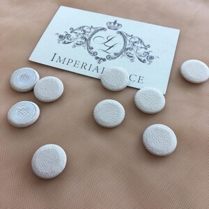 Peut inclure: Plusieurs boutons ronds recouverts de tissu blanc cassé sont disposés sur une surface pêche. Une carte avec le texte "IMPERIAL LACE" et un blason orné est également présente.