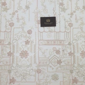 May include: White beaded lace fabric with a geometric pattern and pink floral accents. The fabric is shown on a white background with a ruler measuring 30 inches.