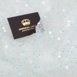 Puede incluir: Tela blanca con pequeñas lentejuelas iridiscentes en un patrón disperso. Las lentejuelas son blancas, doradas y verde azulado. Una pequeña tarjeta negra con el texto "Imperial Lace" y un logotipo de corona es visible en la esquina superior derecha.