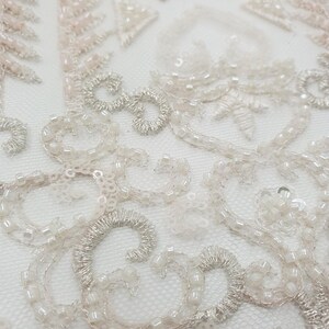 May include: Close-up of delicate white and pink beaded lace fabric with a floral design. The fabric is made of a sheer mesh material and features intricate beading and embroidery.