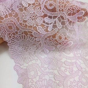 Puede incluir: Primer plano de una delicada tela de encaje de color lila claro. El encaje presenta un intrincado patrón floral con costuras detalladas y un borde festoneado. La tela es semitransparente, revelando la textura y el diseño.