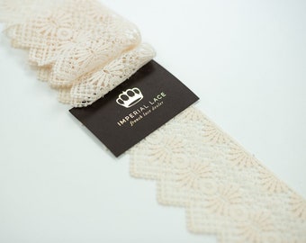 May include: A close-up of a cream-colored lace trim with a scalloped edge. The lace has a delicate floral pattern. A brown tag with gold lettering reads "Imperial Lace French Lace Dealer".