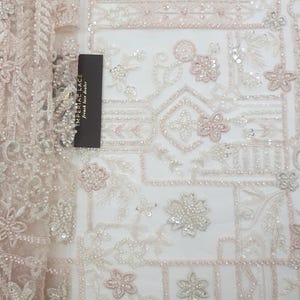 May include: A delicate, beaded lace fabric with a geometric pattern in shades of white and pink. The fabric features intricate floral designs and small sequins.
