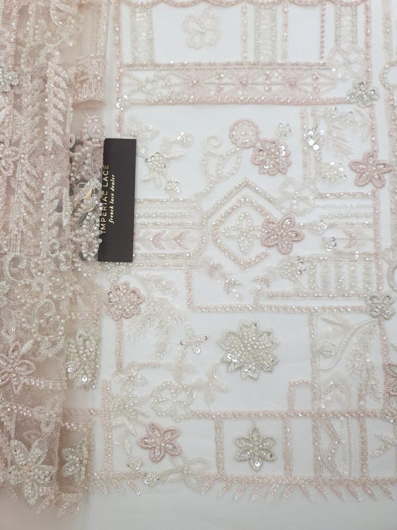 May include: A delicate, beaded lace fabric with a geometric pattern in shades of white and pink. The fabric features intricate floral designs and small sequins.