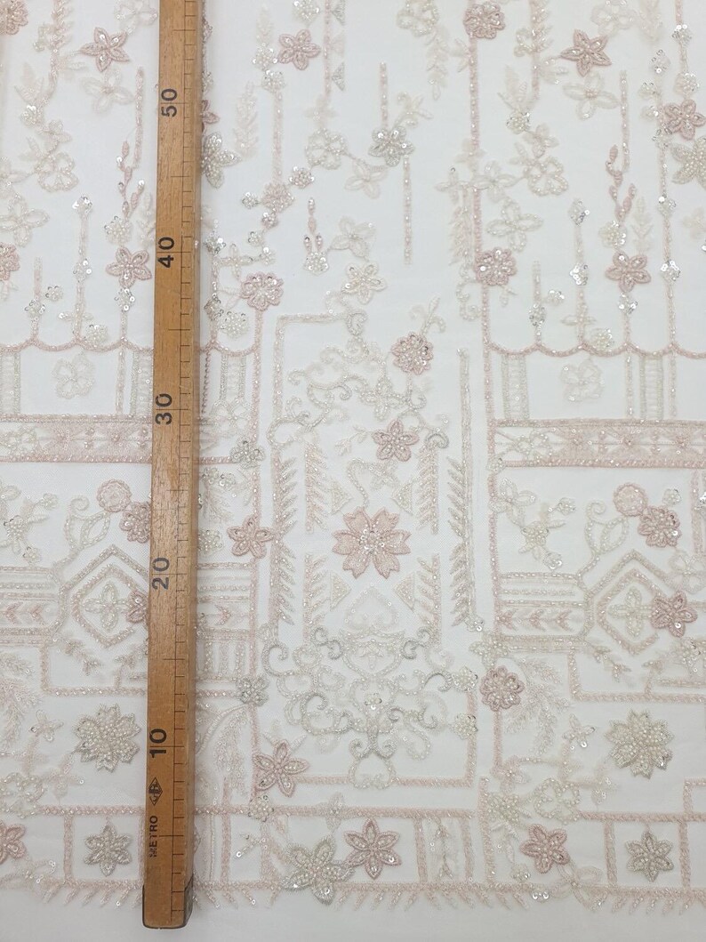 May include: White and pink beaded lace fabric with a geometric pattern. The fabric is shown with a ruler measuring 50 inches.