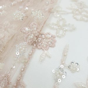 May include: Close-up of delicate blush pink tulle fabric with intricate floral embroidery and white sequins. The fabric is embellished with small white beads and sequins, creating a shimmering effect.
