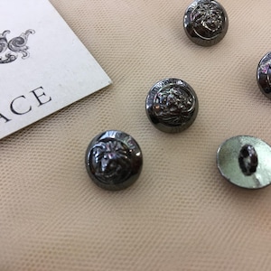 May include: Six silver-toned buttons with a raised design of a lion's head. The buttons are round and have a smooth, shiny finish.