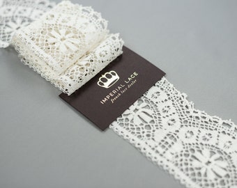May include: White cotton lace trim with a delicate floral pattern. The lace is folded and displayed on a brown card with the text "IMPERIAL LACE French lace dealer".