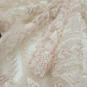 May include: Close-up of delicate, beaded lace fabric in a blush pink and ivory color scheme. The lace features intricate floral patterns and small, iridescent beads.