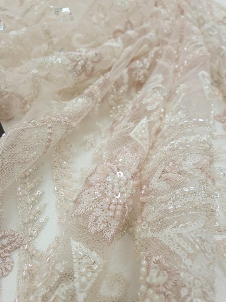 May include: Close-up of delicate, beaded lace fabric in a blush pink and ivory color scheme. The lace features intricate floral patterns and small, iridescent beads.