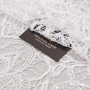 May include: White lace fabric with a delicate floral pattern. The fabric is embellished with small sequins. A brown card with the text "Imperial Lace French Lace Dealer" is partially visible in the bottom right corner of the image.