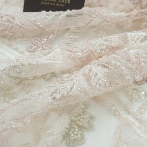 May include: Close-up of delicate, blush-colored lace fabric with intricate floral patterns and beaded embellishments. The fabric is soft and sheer, with a subtle shimmer.