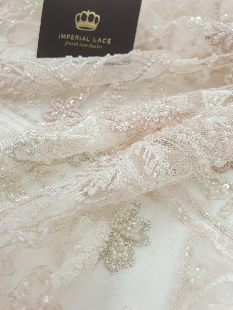 May include: Close-up of delicate, blush-colored lace fabric with intricate floral patterns and beaded embellishments. The fabric is soft and sheer, with a subtle shimmer.