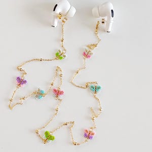 May include: Gold-coloured earbud chains adorned with beaded butterfly charms in shades of pink, green, purple, and blue. The chains are attached to white earbuds. The butterfly charms are made of small beads.