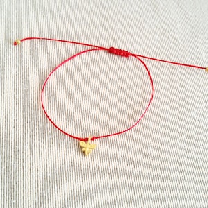 May include: A red string bracelet with a gold butterfly charm. The bracelet is tied with a knot and has gold beads at the ends.