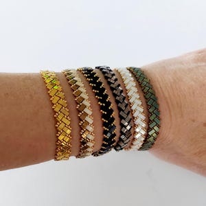 May include: Six beaded bracelets in various colors, including gold, cream, black, gray, white, and green. The bracelets have a woven design and are displayed on a person's arm. The bracelets are a fashion accessory.