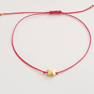 May include: A red string bracelet with a gold-coloured butterfly charm. The bracelet is adjustable with a sliding knot closure and small gold beads at the ends. The butterfly charm is in the centre of the red string.