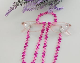 May include: Pink and fuchsia beaded glasses chain with flower-shaped beads. The chain is looped around a pair of pink glasses.