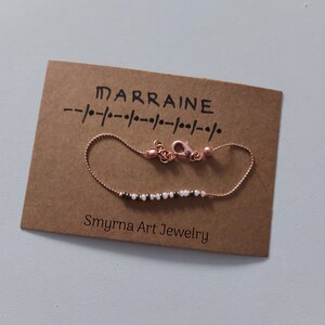 May include: A delicate gold chain bracelet with small white and black beads. The bracelet is displayed on a brown card with the text "MARRAINE" and "Smyrna Art Jewelry".