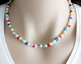 May include: A delicate pearl necklace with colorful accents. The necklace features small, round white pearls interspersed with vibrant red, blue, green, and yellow beads and flower-shaped charms. The necklace is displayed on a mannequin.