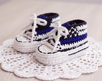 May include: A pair of handmade crochet baby sneakers. The shoes are white with black and blue accents, including the laces and striped design. The sneakers are displayed on a white, lacy doily.