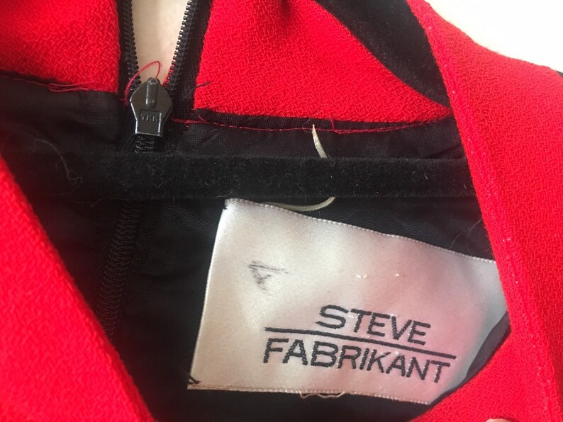 May include: Close-up of a black and red dress with a zipper and a white label that reads "STEVE FABRIKANT".